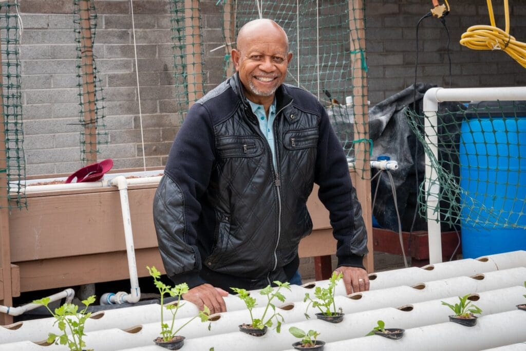 Photo shows Dr. George Brooks, an African American man, standing behind plants being grown using aquaponics.